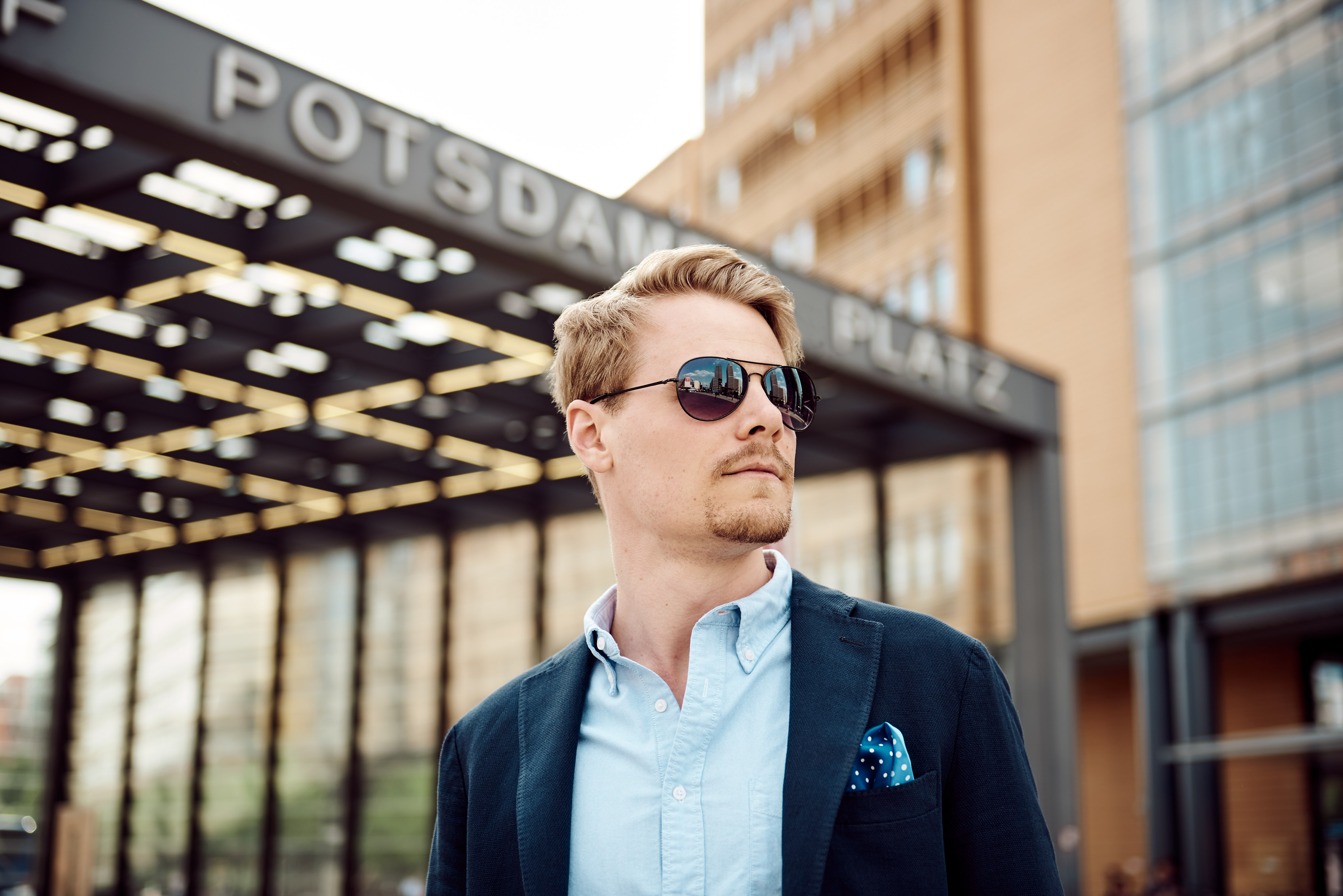ein junger Mann mit Sportsakko, Sonnenbrille und blondem Bart schaut am Potsdamer Platz in Berlin an der Kamera vorbei
