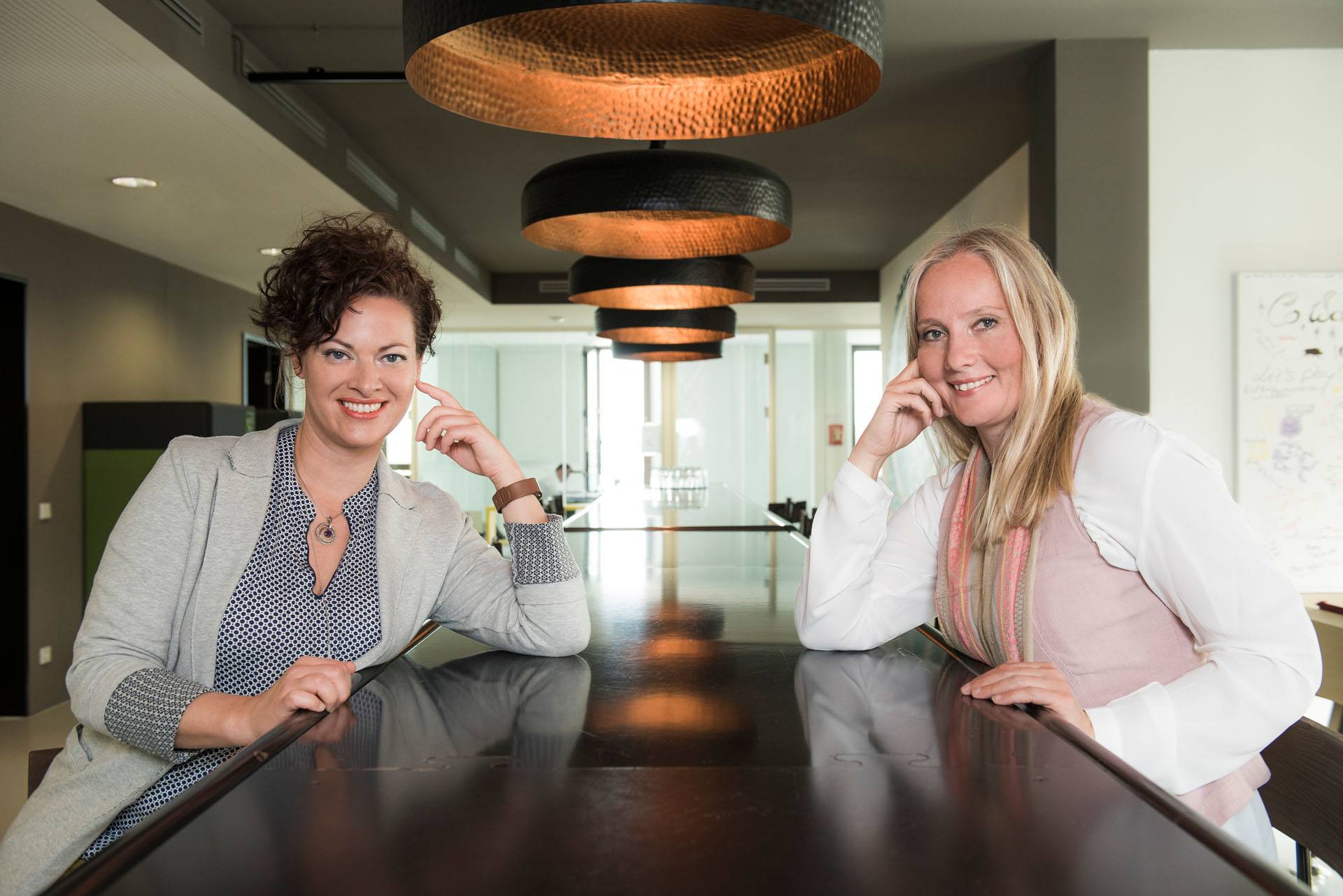 zwei weibliche Coaches und Trainerinnen sitzen an einem Tisch eines modernen Coworking-Spaces und lächeln in die Kamera